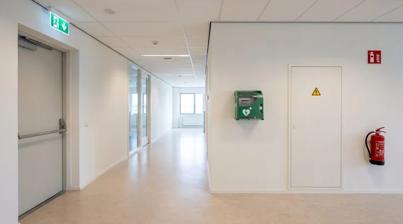 Een lichte gang met een nooduitgangbord, een AED-kast en een brandblusser aan de muur